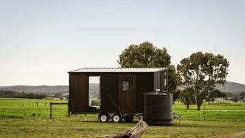 A small, dark-colored cabin with a sliding door, a water tank, and a trailer attached.