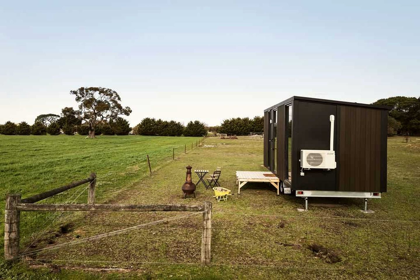 A small, dark-colored mobile home with a white air conditioning unit, situated in a grassy field with a wooden fence in the foreground.