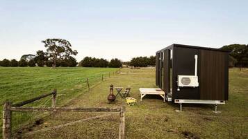 A small, dark-colored mobile home with a white air conditioning unit, situated in a grassy field with a wooden fence in the foreground.