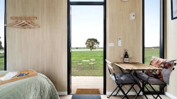 Een slaapkamer met een bed, een houten tafel met een fles en glazen, en uitzicht op een grasveld door een groot raam.