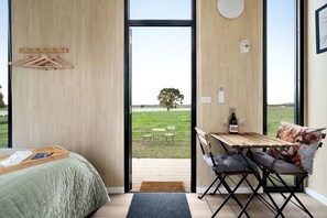 A bedroom with a bed, a wooden table with a bottle and glasses, and a view of a grassy field through a large window.