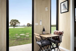 A room with a wooden table and chairs, a bottle of wine, and a view of a grassy field with trees.
