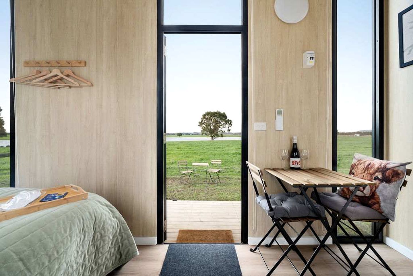 A bedroom with a bed, a wooden table with a bottle and glasses, and a view of a grassy field through a large window.