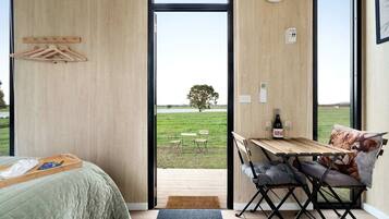 A bedroom with a bed, a wooden table with a bottle and glasses, and a view of a grassy field through a large window.