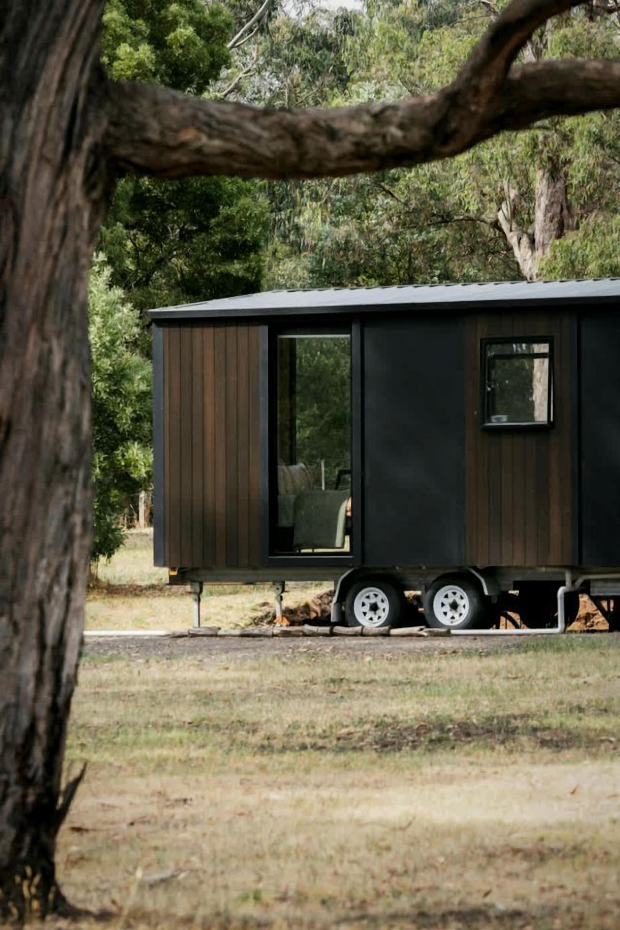 A stationary, dark-colored, single-story mobile home with a sliding door and a window, situated in a grassy area with trees in the background.