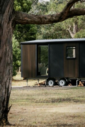A stationary, dark-colored, single-story mobile home with a sliding door and a window, situated in a grassy area with trees in the background. - Delightfully Tiny House Ideal for a Peaceful Escape in Springmount, Victoria (Creswick)