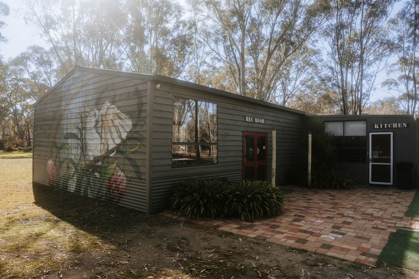 A rustic building with a 'REC ROOM' and 'KITCHEN' labeled, a mural on the side, and a paved entrance area.