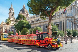 A red and yellow tram with a vintage design, traveling on a street with historical buildings and a large tree in the background. - Vrbo Property (Catania)