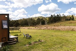 Eine Holzhütte mit einer Terrasse, zwei Stühlen und einem Tisch in einem grünen Feld, im Hintergrund Bäume.