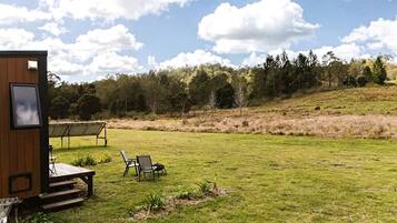 Eine Holzhütte mit einer Terrasse, zwei Stühlen und einem Tisch in einem grünen Feld, im Hintergrund Bäume.