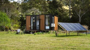 Eine moderne, kompakte Hütte mit einer Terrasse, umgeben von üppiger Vegetation und einer Anlage von Solarmodulen.
