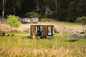Una cabana petita i moderna amb façana de fusta, situada en un prat amb un panell solar i una tanca metàl·lica al fons.