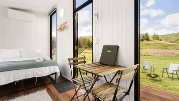 A bedroom with a bed, a desk, and a laptop. There is a view of a grassy outdoor area through a large window.