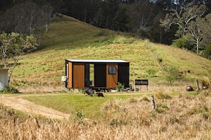 A small cabin with a single door and window, situated on a grassy hill with a dirt path leading to it.