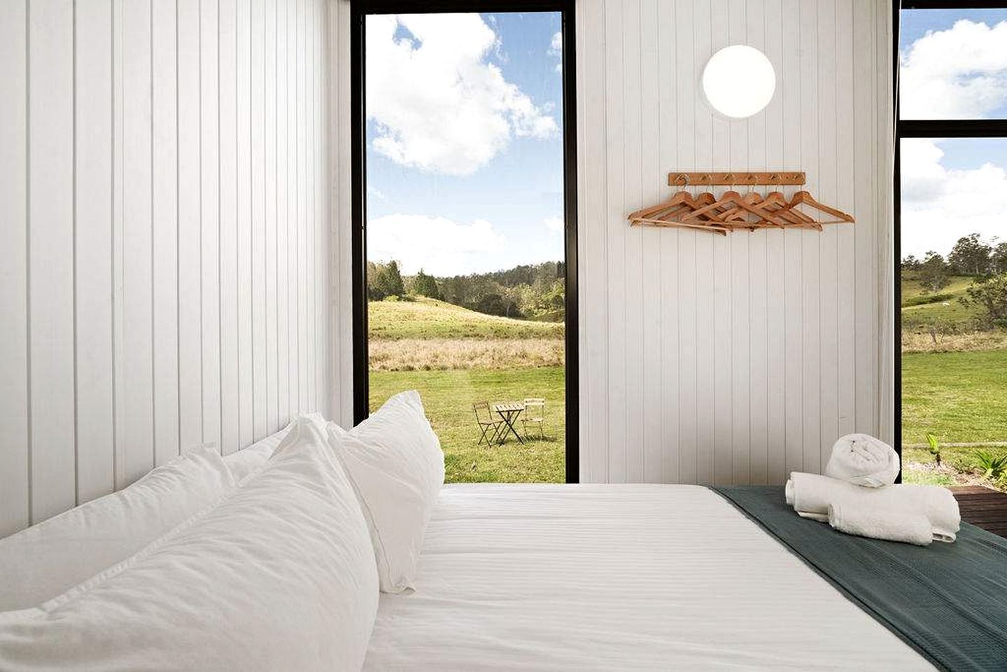 A bedroom with a large window, a bed with white linens, a hanging wooden rack, and a view of a grassy landscape.