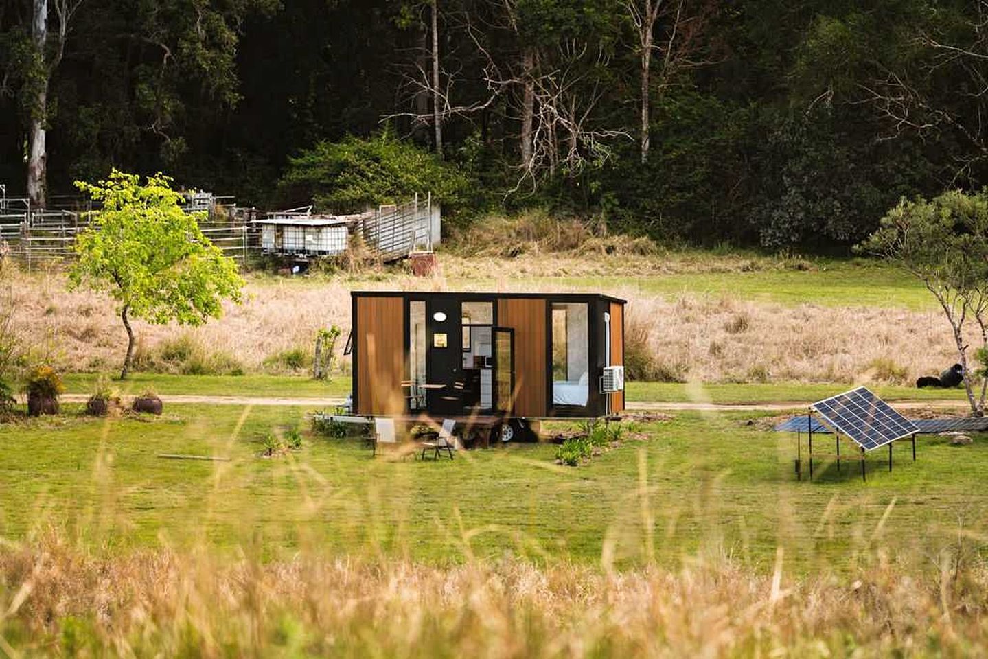 Une petite cabane moderne, aux murs en bois, située dans un champ verdoyant. On aperçoit un panneau solaire et une clôture en métal à l’arrière-plan.
