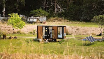 Une petite cabane moderne, aux murs en bois, située dans un champ verdoyant. On aperçoit un panneau solaire et une clôture en métal à l’arrière-plan.