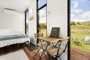 A bedroom with a bed, a desk, and a laptop. There is a view of a grassy outdoor area through a large window.