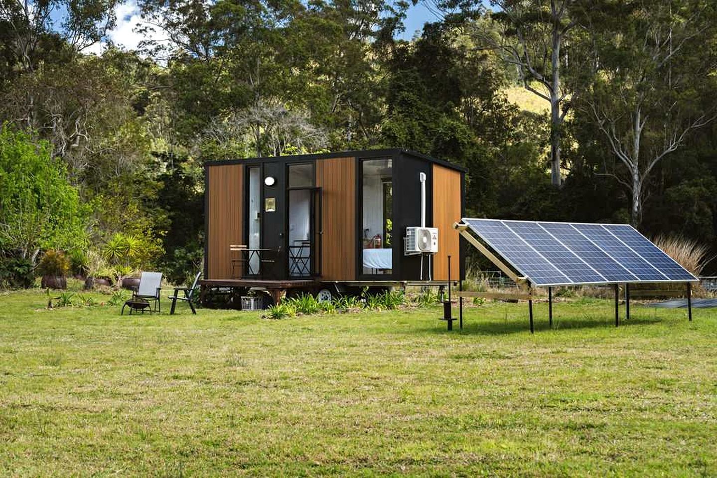 A modern, compact cabin with a deck, surrounded by lush greenery and a solar panel array.