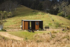 Une petite cabane avec une seule porte et une seule fenêtre, située sur une colline herbeuse, accessible par un sentier de terre.
