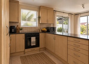 A kitchen with wooden cabinets, a stove, a microwave, and a window with curtains. - Gorgeous 3-bedroom apartment in Muizenberg. (Cape Town)