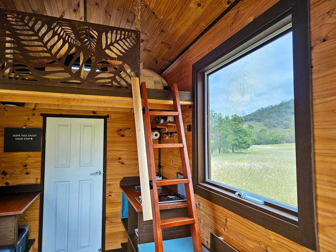 A wooden cabin interior with a ladder, a white door, and a window overlooking a grassy field.