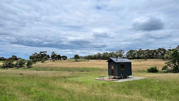 Une petite cabane de couleur sombre, dotée d’une seule porte et d’une seule fenêtre, située dans un champ herbeux, avec des arbres en arrière-plan.