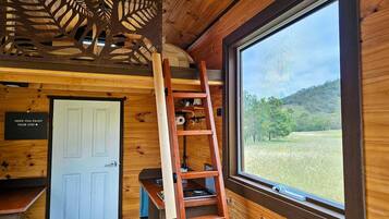 A wooden cabin interior with a ladder, a white door, and a window overlooking a grassy field.