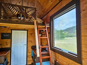A wooden cabin interior with a ladder, a white door, and a window overlooking a grassy field.