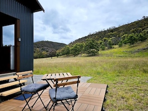 An outdoor wooden deck with a table and chairs, overlooking a grassy field and hills.