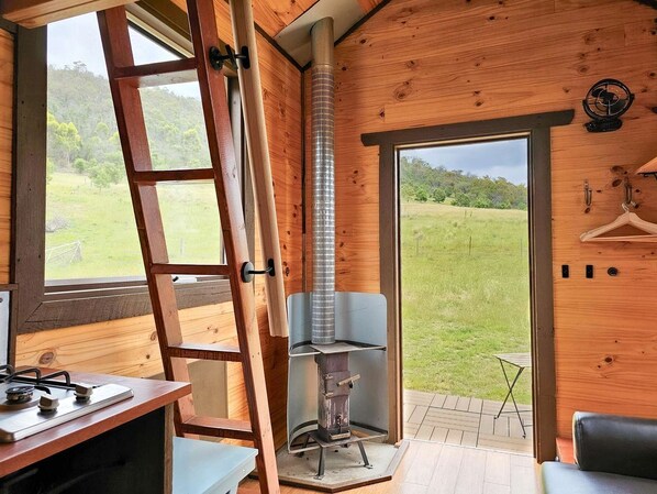 A wooden interior with a stove, a ladder, and a view of the outdoors.