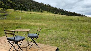 An outdoor seating area with a table and two chairs on a wooden deck, overlooking a grassy field and a forested hill.