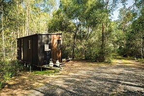 Uma RV pequena e escura, com aquecedor de água e tanque de propano, estacionada em um caminho de terra cercada por mata densa.