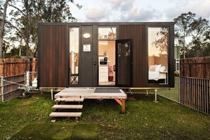 A compact cabin with a sliding door entrance, a small outdoor seating area, and a visible kitchen area. - Lovely Tiny House in Wongawallan, Queensland (Wongawallan)