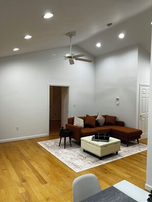 A spacious living room with a brown sectional sofa, a white ottoman, a small side table, and a ceiling fan. - The Brook House - Private Woodland Retreat in the Berkshires (Chester)