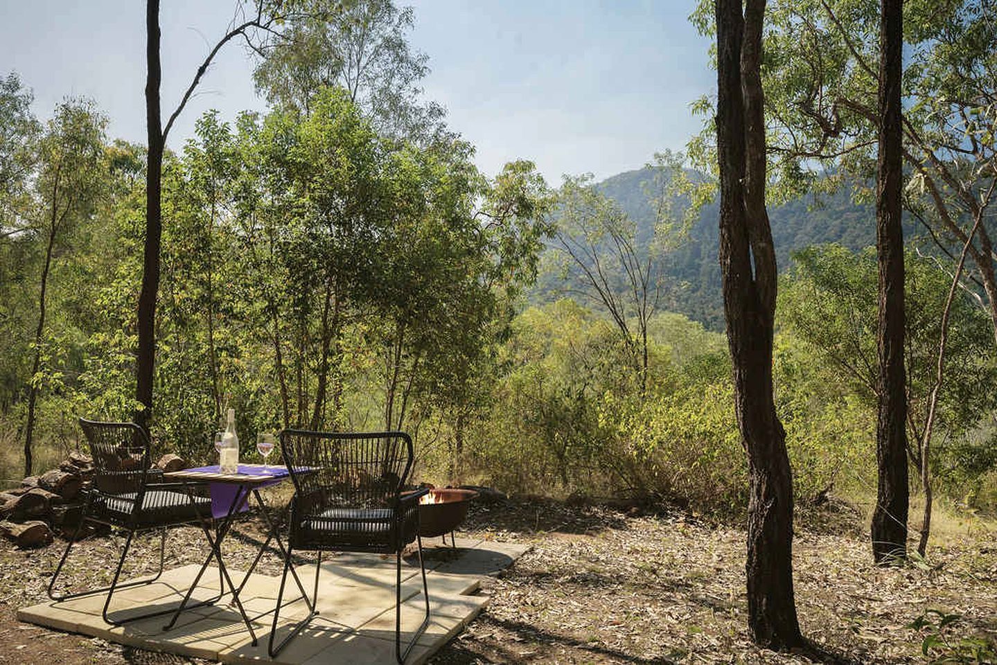 Un área de comedor al aire libre con sillas y mesa de metal, rodeada de árboles y montañas al fondo.