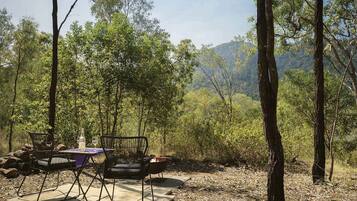 Un área de comedor al aire libre con sillas y mesa de metal, rodeada de árboles y montañas al fondo.