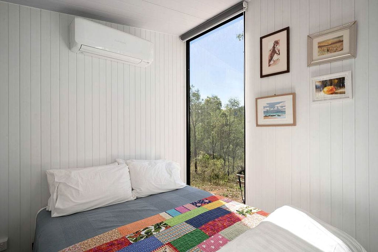 Un dormitorio con una cama, un colcha colorida, una ventana con vista a árboles y cuadros en la pared.