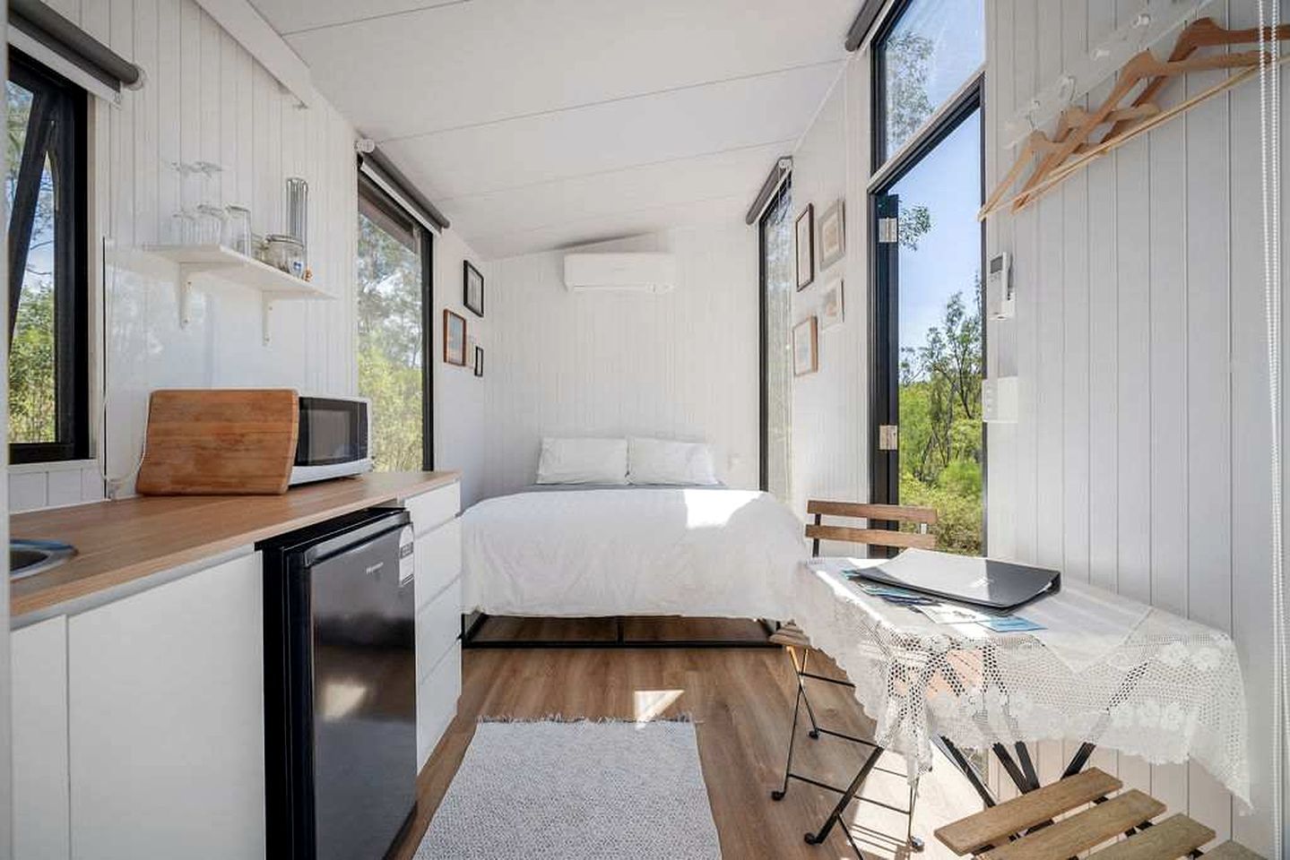 A compact bedroom with a bed, bedside table, and a view of greenery through large windows.