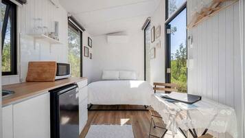 A compact bedroom with a bed, bedside table, and a view of greenery through large windows.