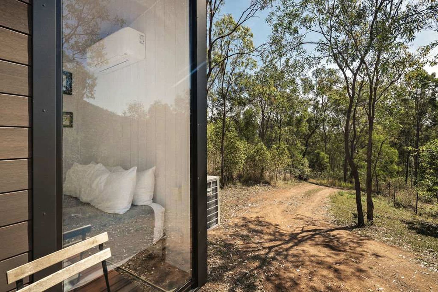 Un bâtiment moderne doté d’une porte en verre, un lit avec une literie blanche et une vue sur un chemin de terre qui traverse une forêt.