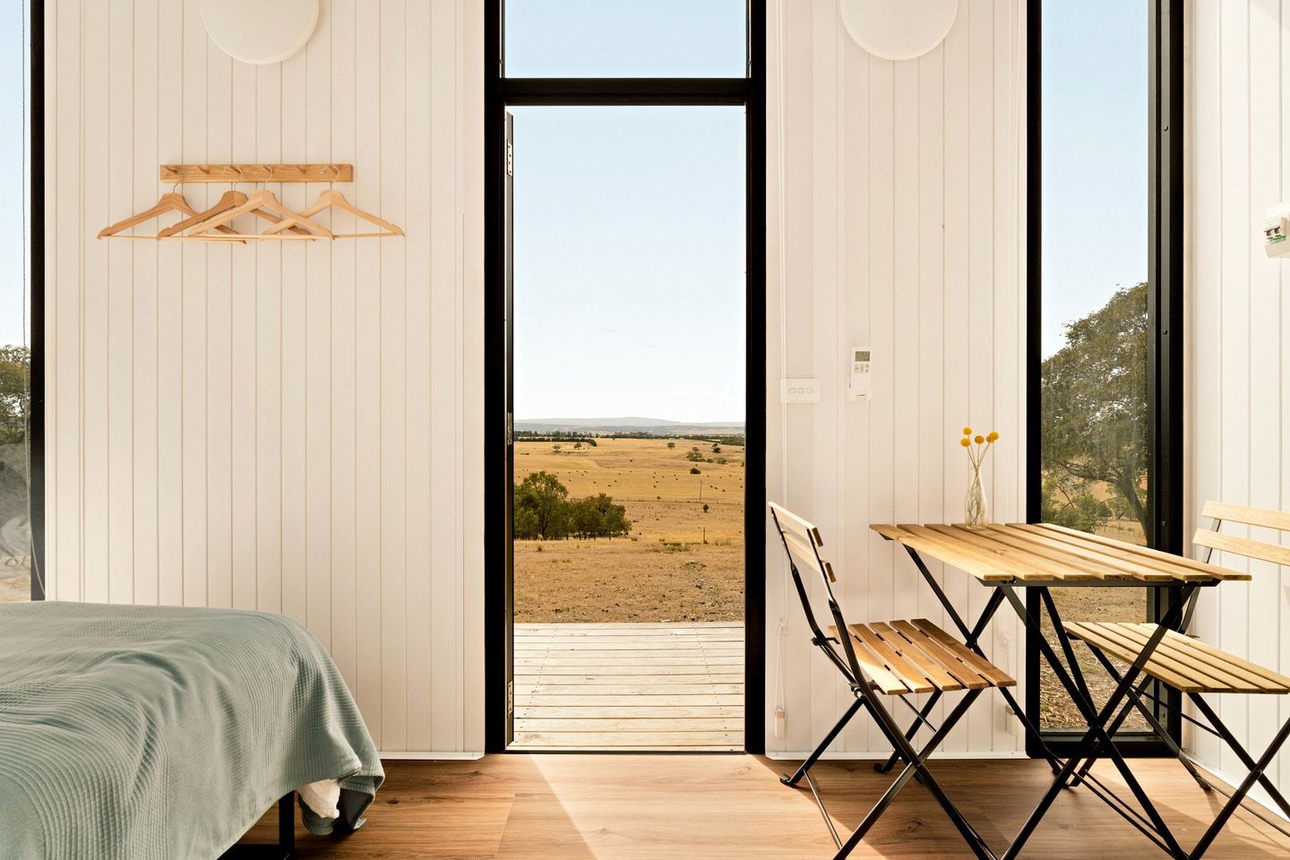 A bedroom with a bed, a wooden table with chairs, and a view of a field through a large window.