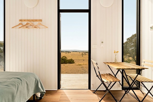 A bedroom with a bed, a wooden table with chairs, and a view of a field through a large window.