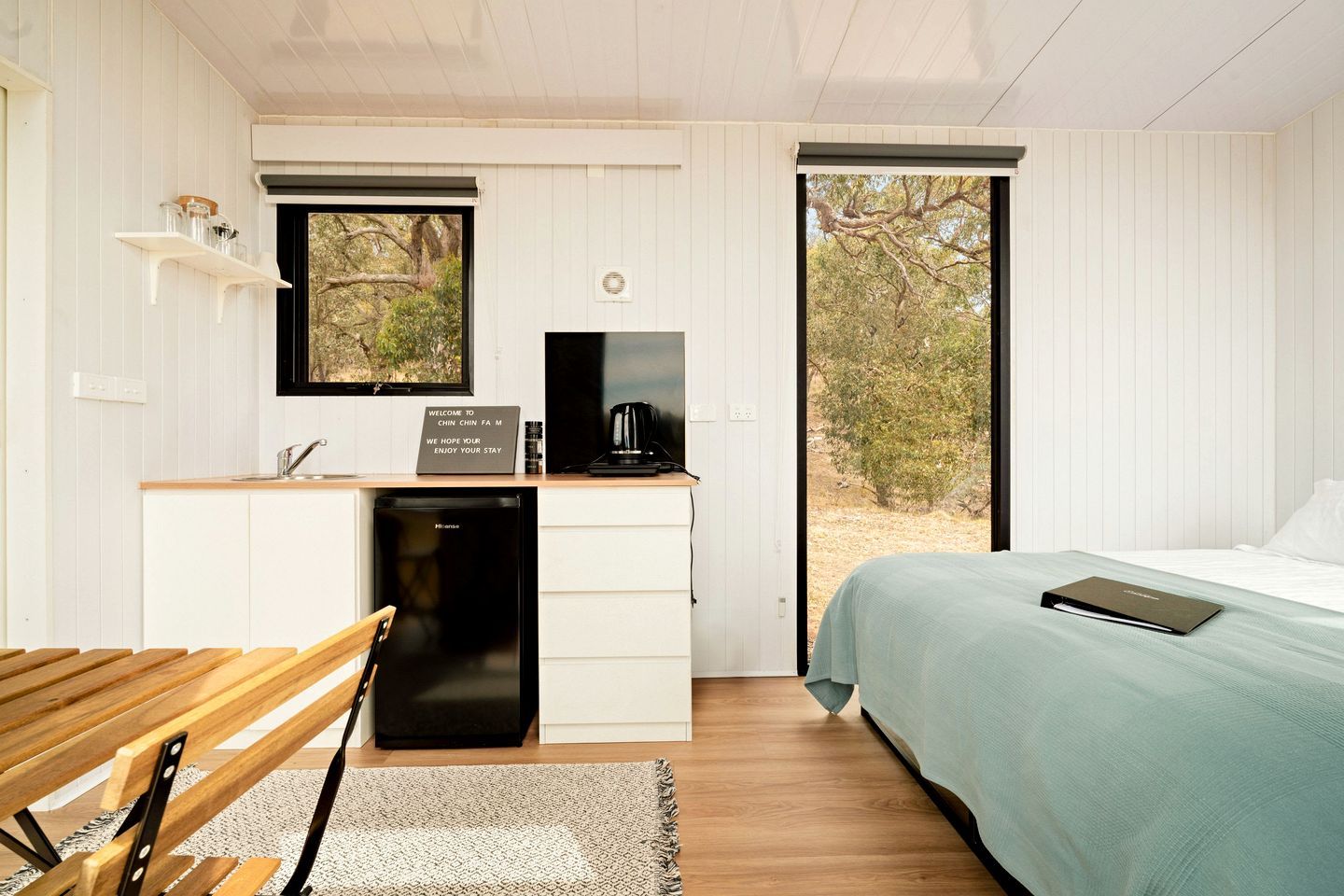 A compact room with a bed, a desk with a coffee maker, a small sink, and a window with a view of trees.