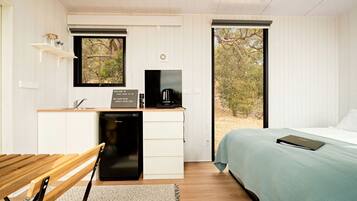 A compact room with a bed, a desk with a coffee maker, a small sink, and a window with a view of trees.