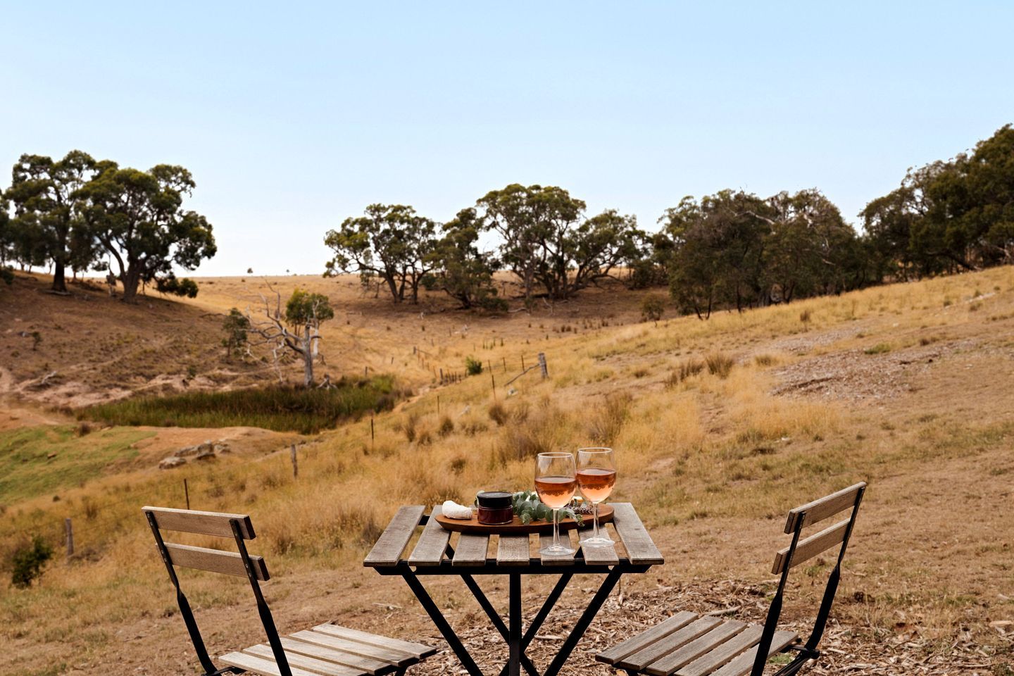 A picnic table with two chairs, two wine glasses, and a small bowl on a grassy hill with trees in the background.