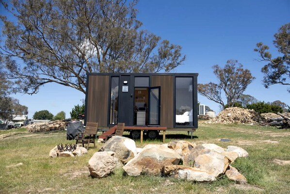 Une cabane moderne et compacte, dotée d’une façade en bois et d’une porte en verre, située dans une zone herbeuse, avec des rochers et des arbres en arrière-plan.