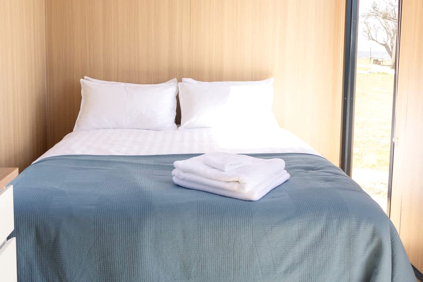 A neatly made bed with a blue bedspread and white pillows, a folded white towel, and a wooden headboard.