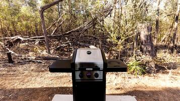 A portable barbecue grill with control knobs and a digital display, set up on a white platform in a forested area.