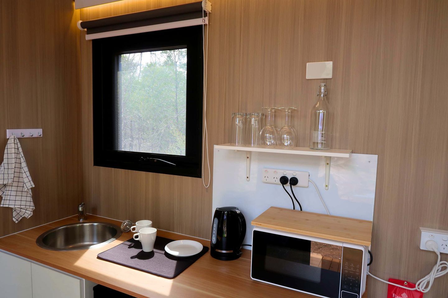 A compact kitchenette with a window, a sink, a microwave, and a shelf with glassware.
